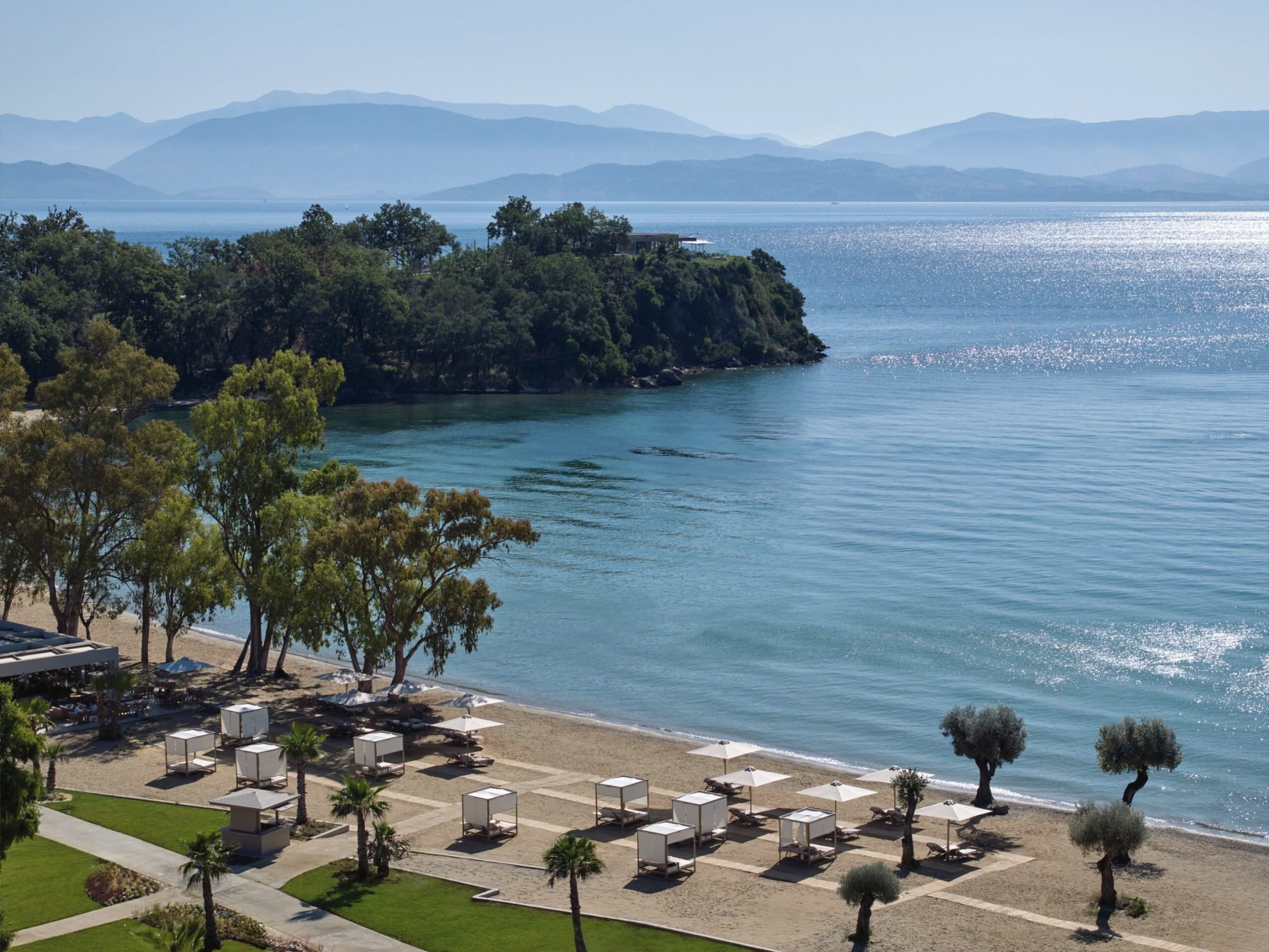 Ikos Odisia's beach cabanas and parasols beneath the trees, on a calm bay with a wooded headland and the mountains of the mainland beyond