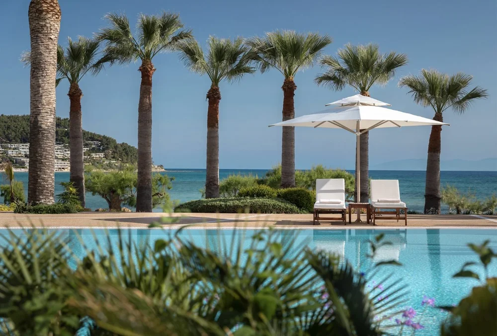 A row of palm trees and white sun loungers beside the pool at Sani Beach, with the sea behind