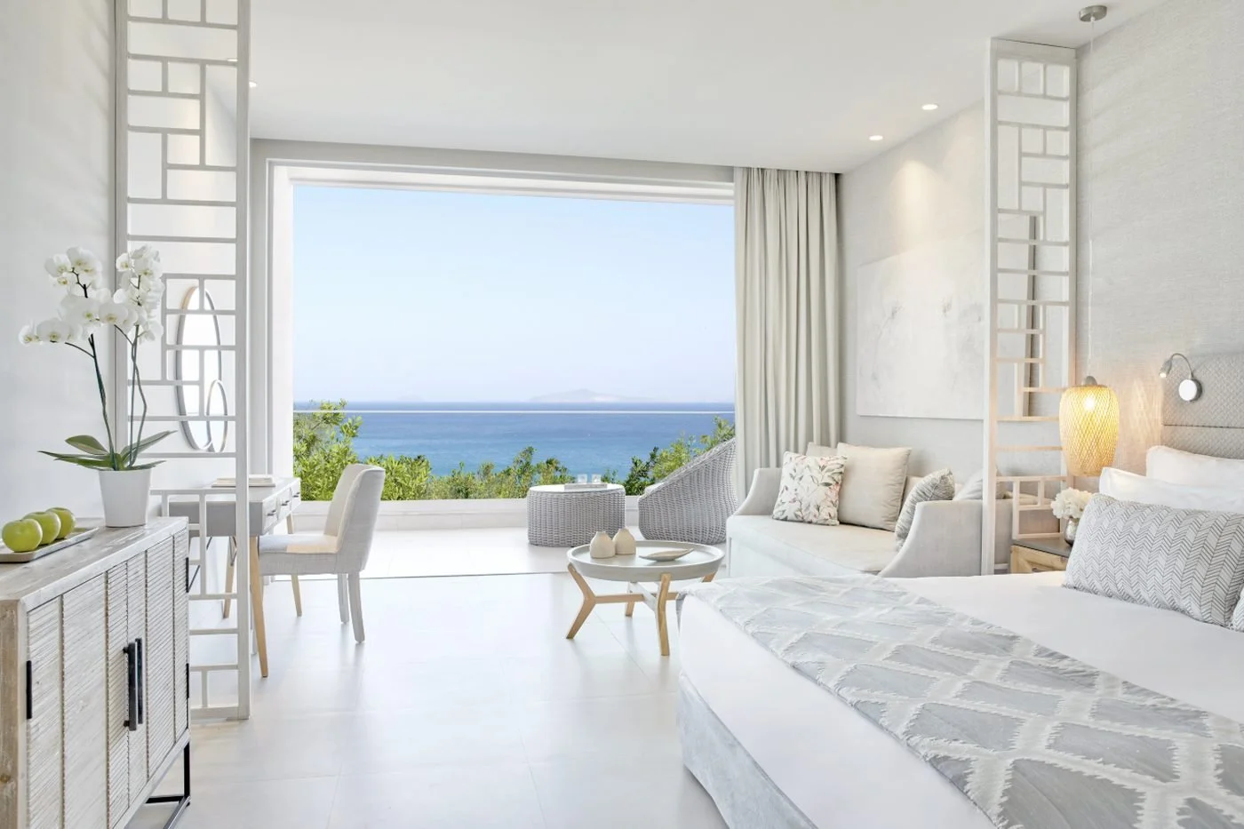 A Junior Suite at Ikos Aria with a bed, sofa and lattice screens, opening onto a balcony with sea views
