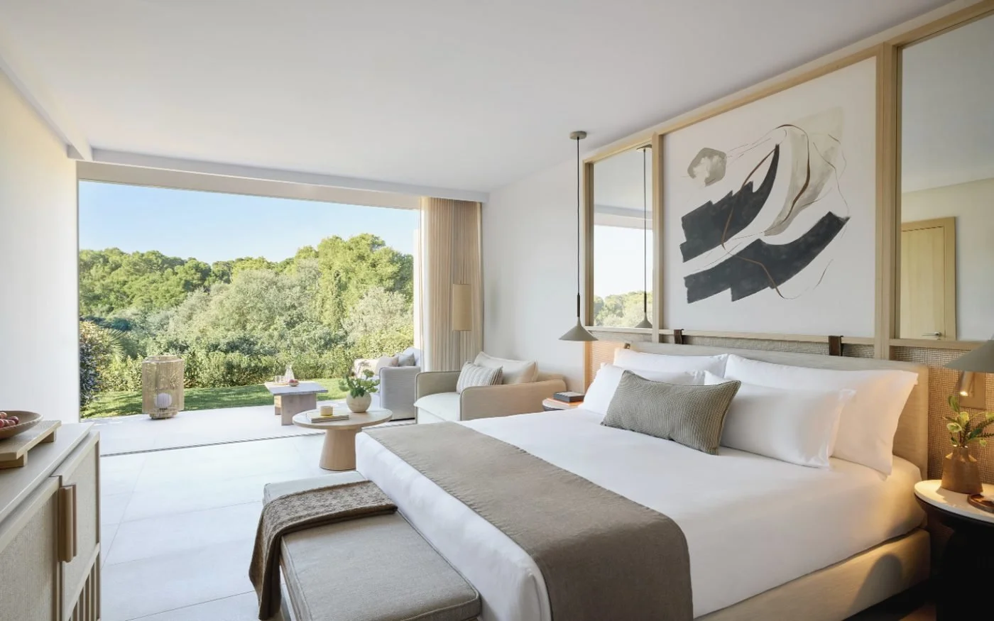A Double Room at Ikos Porto Petro, its bed facing floor-to-ceiling windows, a garden terrace and pine trees