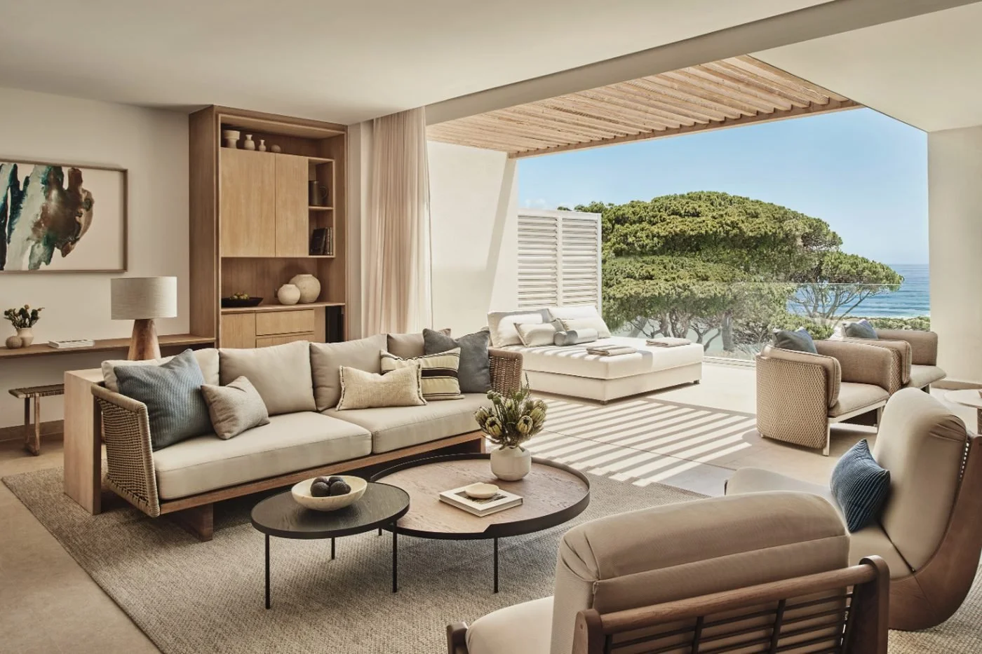 The living room of a Deluxe Two Bedroom Penthouse Suite opening onto a shaded terrace with a daybed, pine trees and the sea