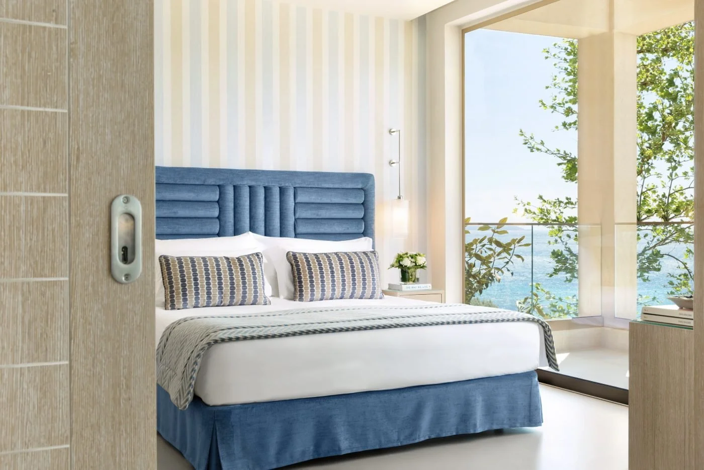 The bedroom of a Deluxe One Bedroom Family Suite at Ikos Oceania, seen through a sliding door, with a balcony over the sea