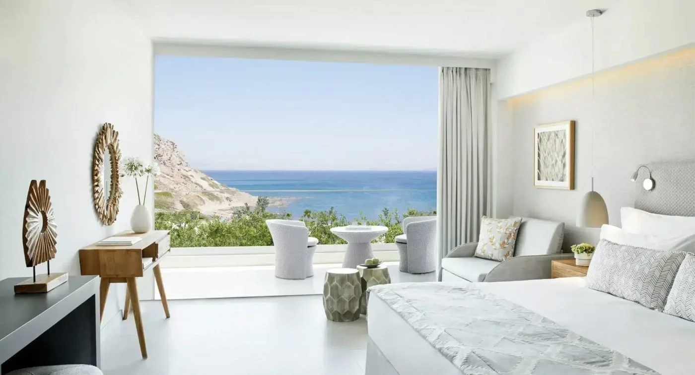 A Superior Triple Room at Ikos Aria with a balcony looking out over greenery to a rocky headland and the sea
