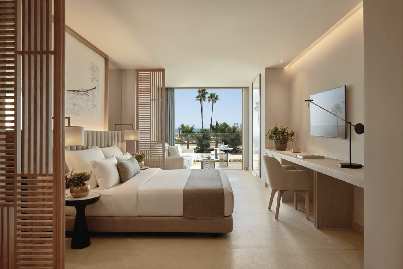 A Junior Suite Pool View with a double bed, a long desk and a balcony looking over palm trees to the pool