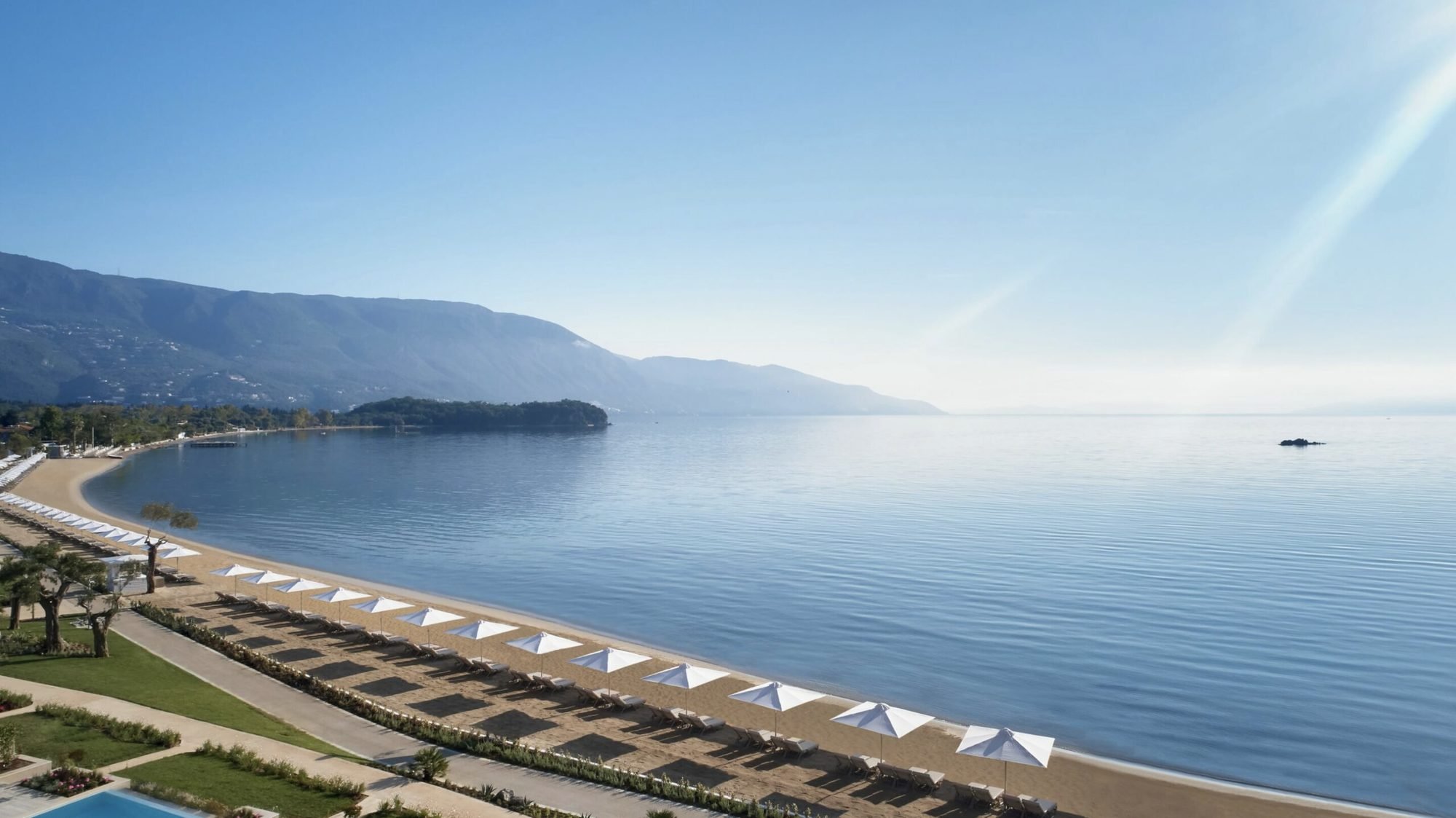 The long curve of Ikos Dassia's beach, lined with white parasols and loungers, on a calm morning with Corfu's mountains beyond