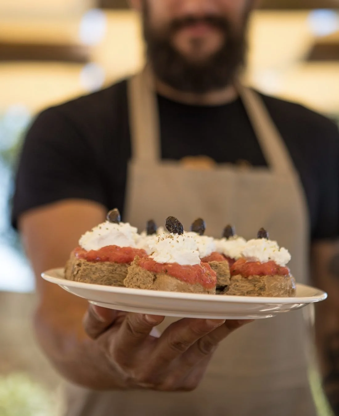 A waiter holding out a plate of Cretan dakos topped with tomato, soft cheese and olives