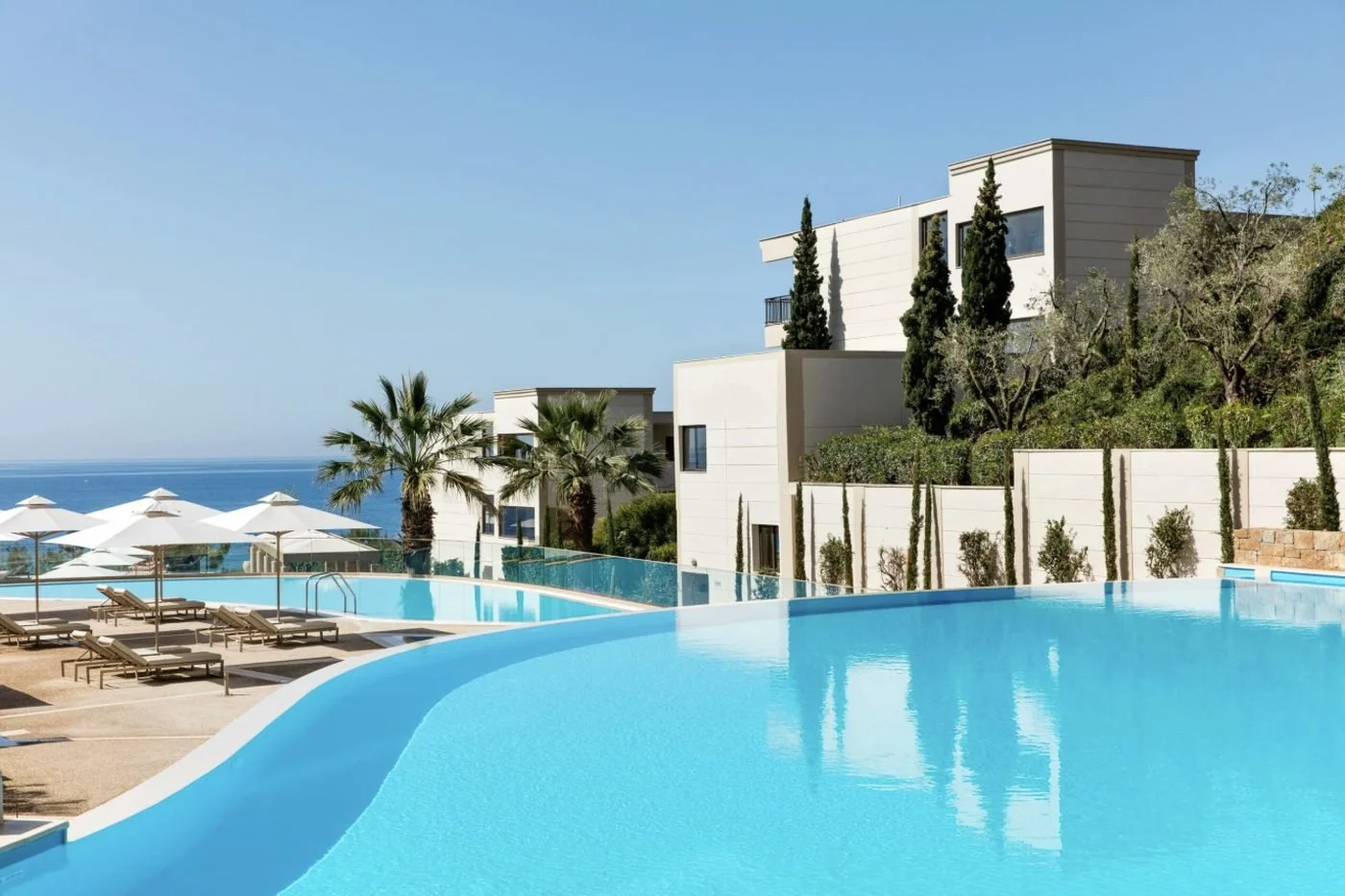 An infinity pool beside white buildings, palms and cypress trees at Ikos Oceania, with the sea beyond