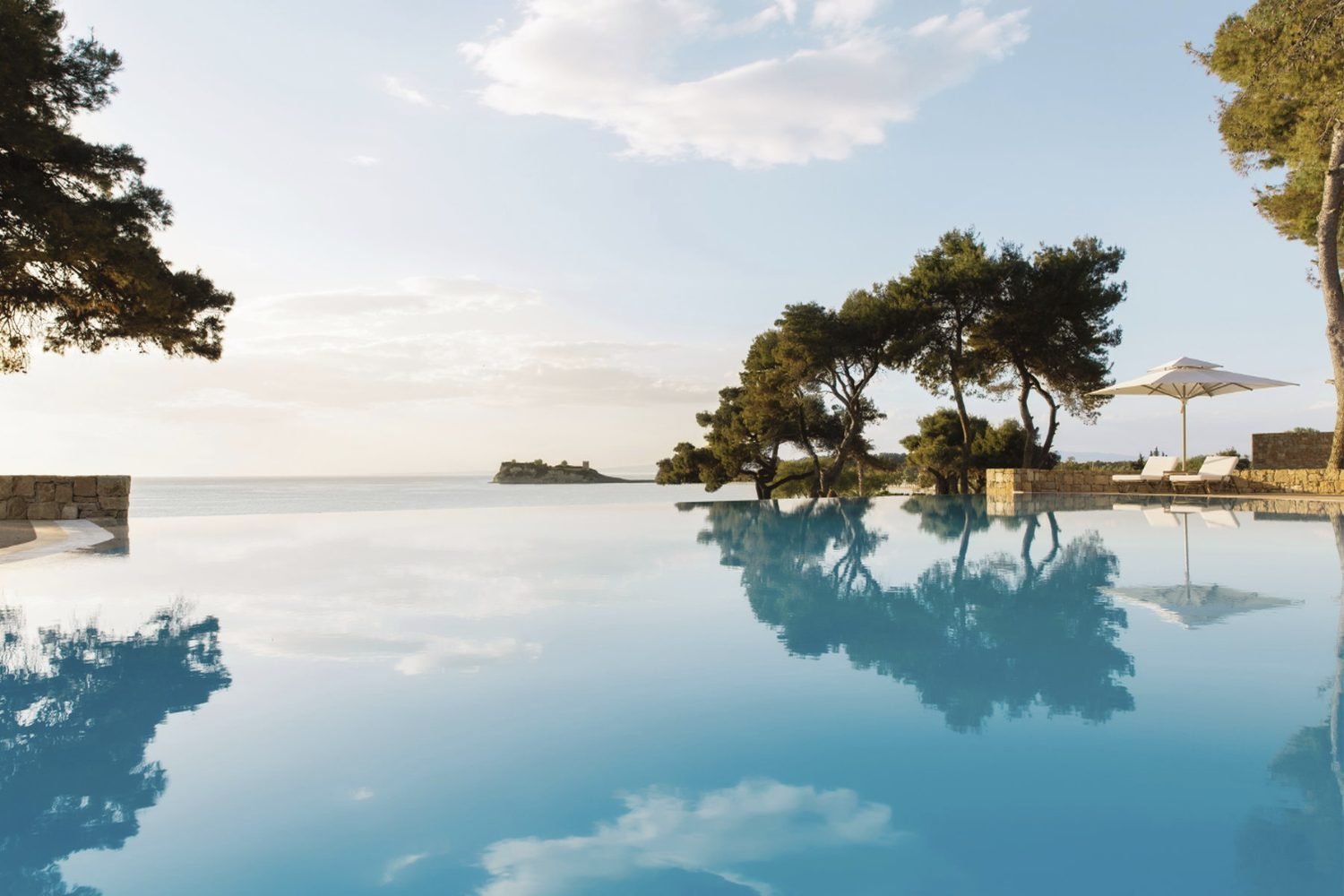 The still infinity pool at Sani Club reflecting pine trees and the sky, with the sea and a headland beyond