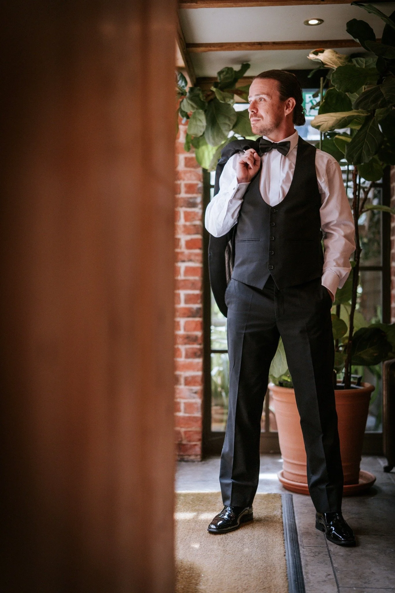 Just Saul in a waistcoat and bow tie, holding his jacket over his shoulder beside leafy plants