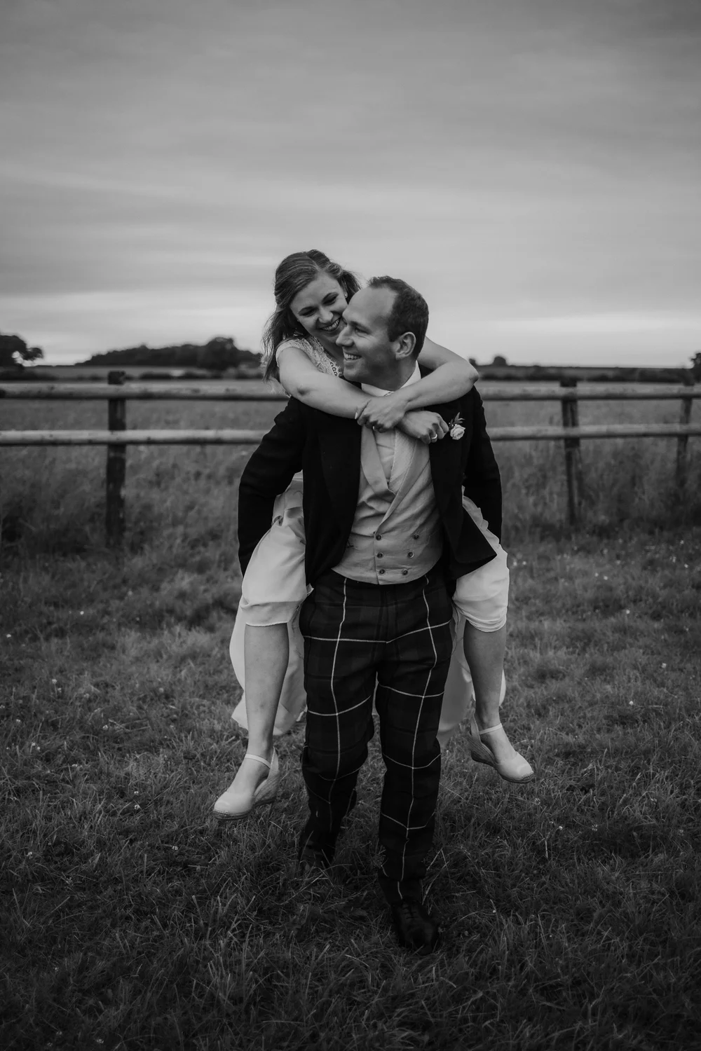 Black and white photo of the groom giving the bride a piggyback across a field