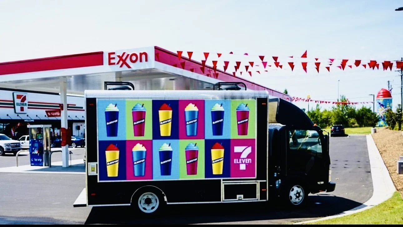 Mobile billboard truck with colorful frozen-drink artwork parked at a convenience store grand opening