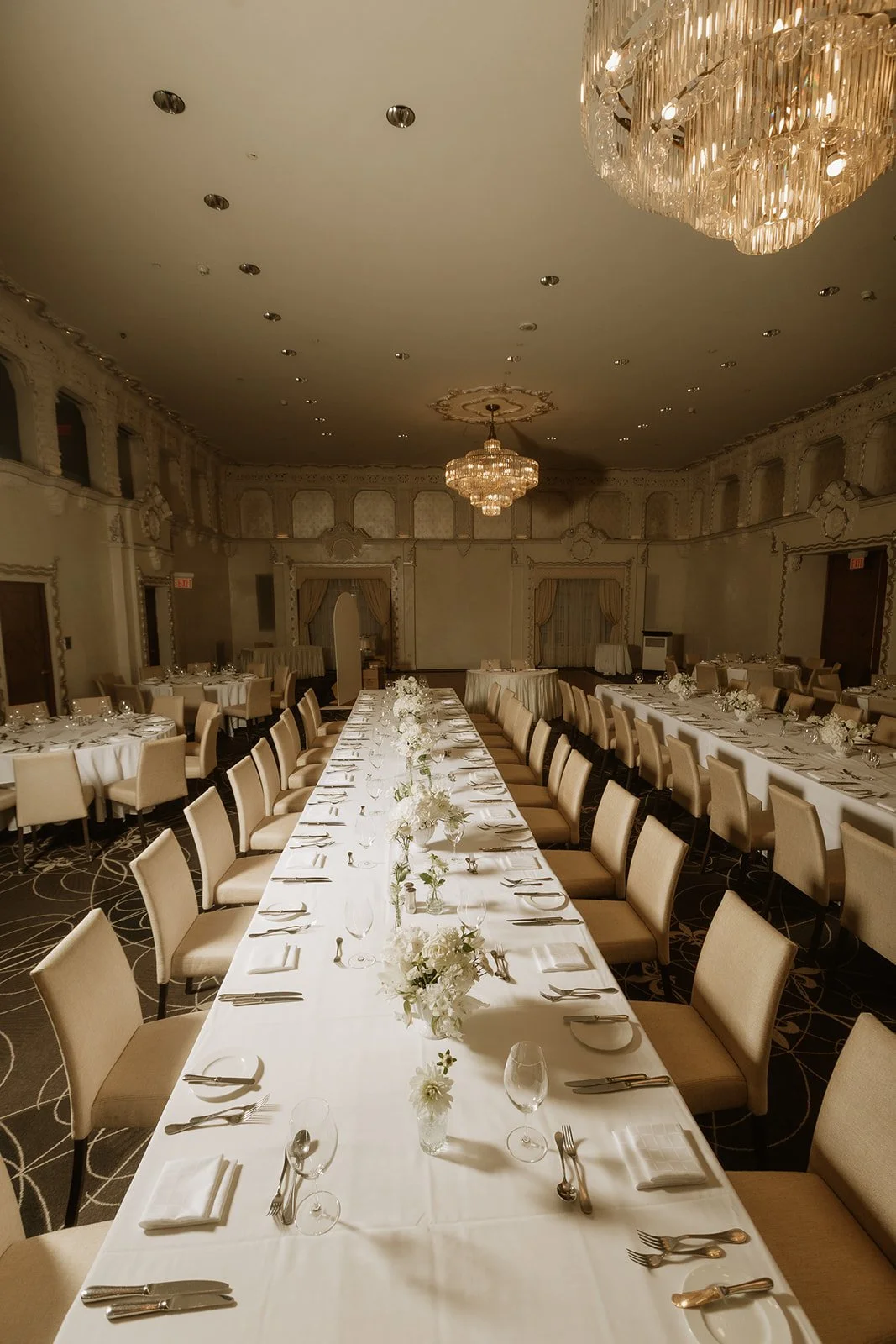 A long dinner table set beneath crystal chandeliers in a cream ballroom