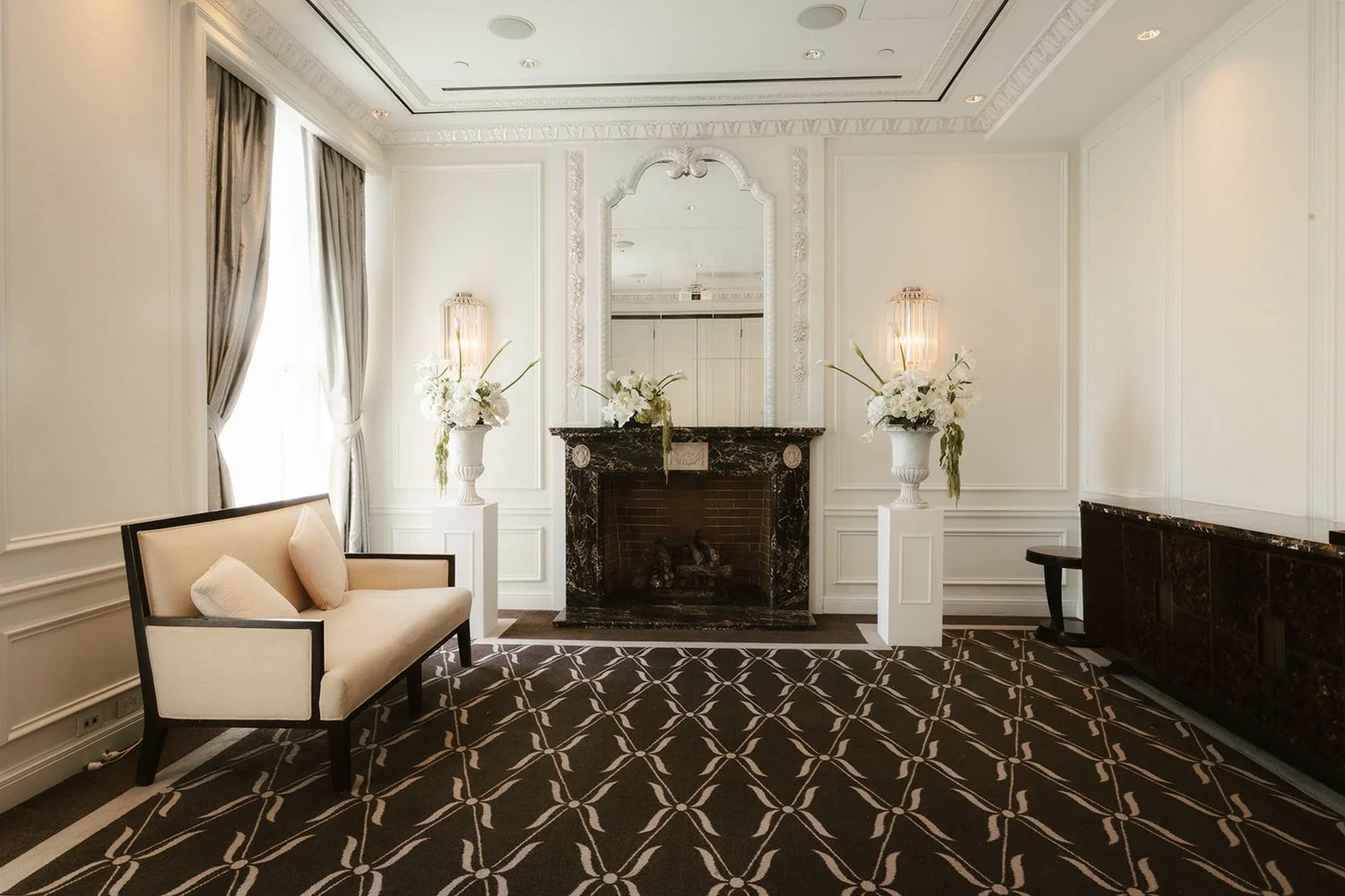 A hotel salon with a marble fireplace, white flowers and a patterned carpet