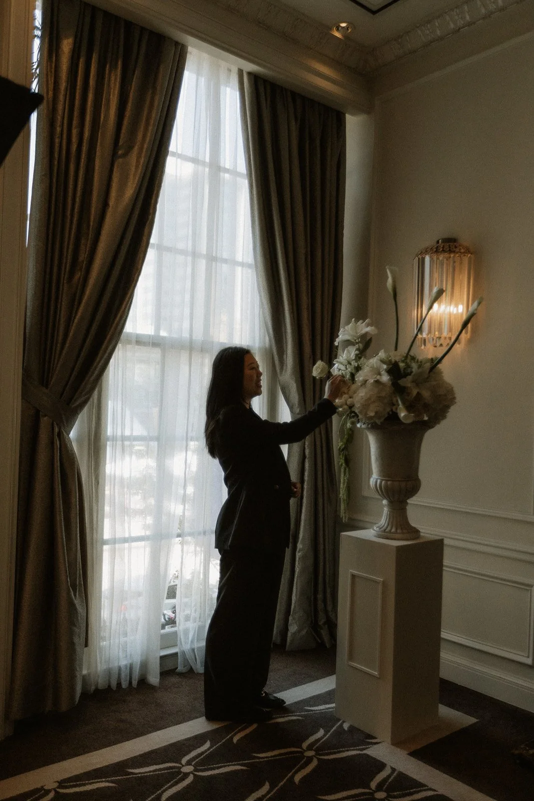 A florist placing white flowers on a pedestal beside tall windows with grey drapes