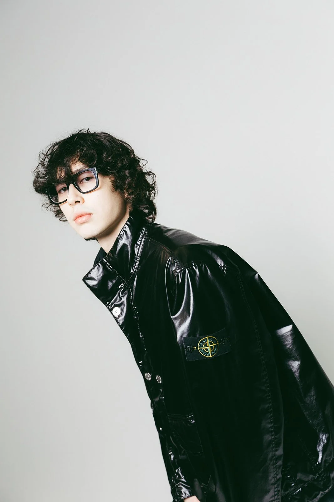 A young man with curly hair and glasses in a glossy black jacket against white