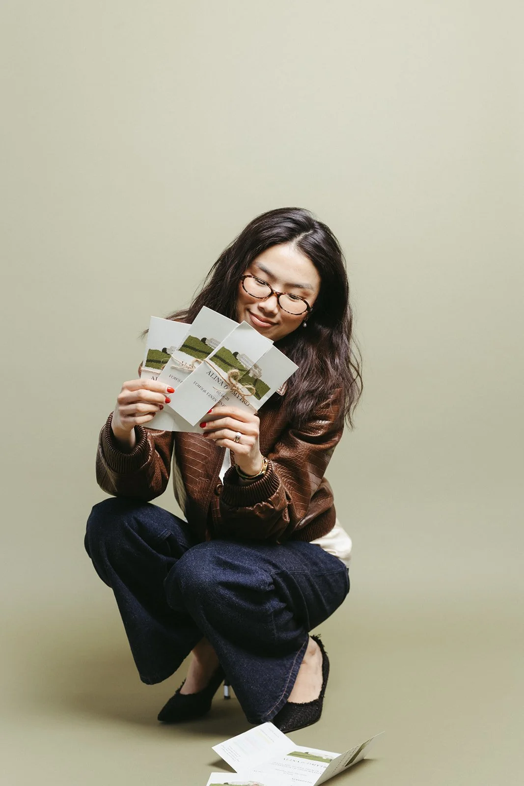 A woman crouching on an olive backdrop, smiling behind a fan of stationery cards