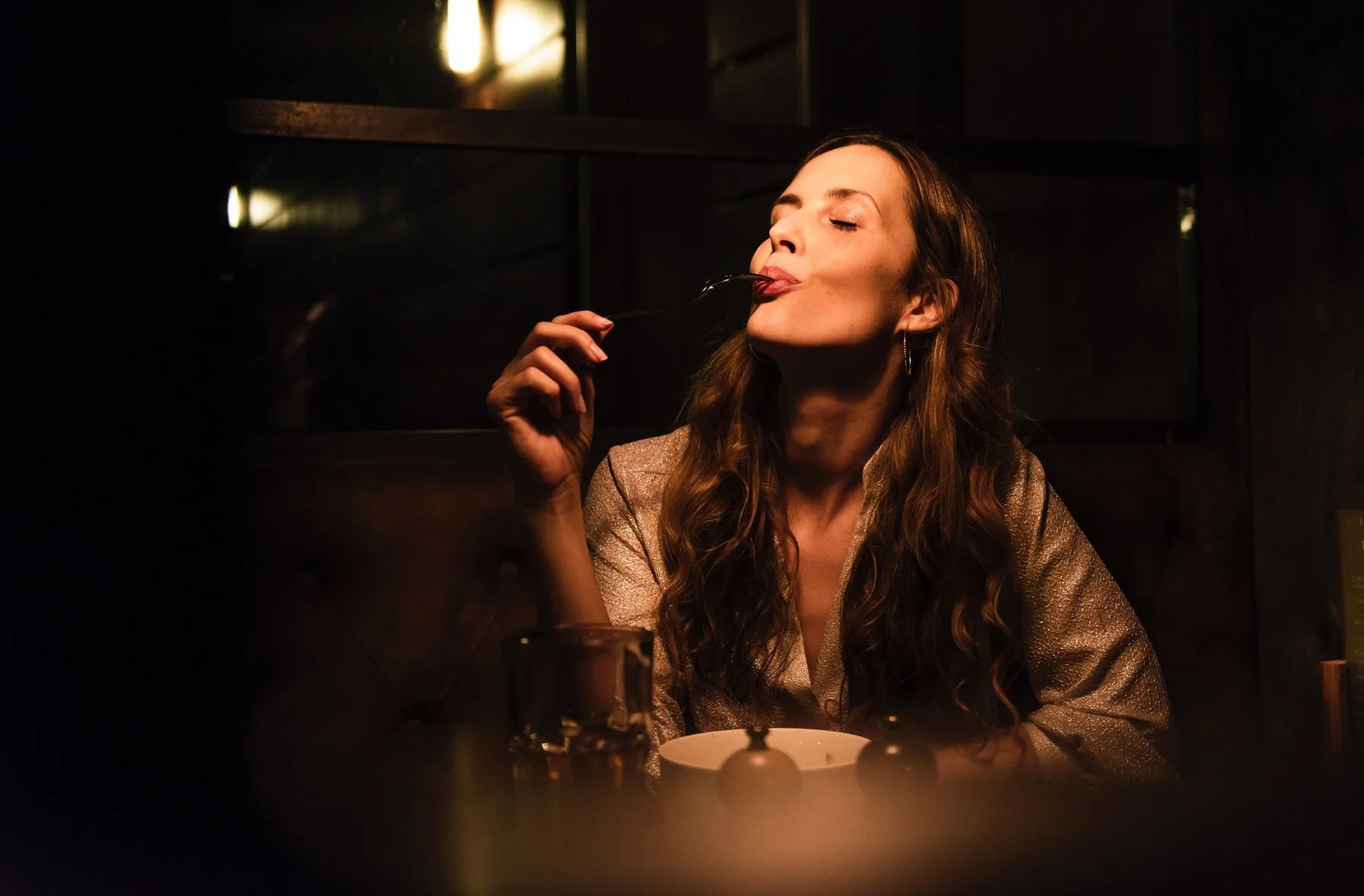 A woman in a dim, warm restaurant licks dessert from her spoon