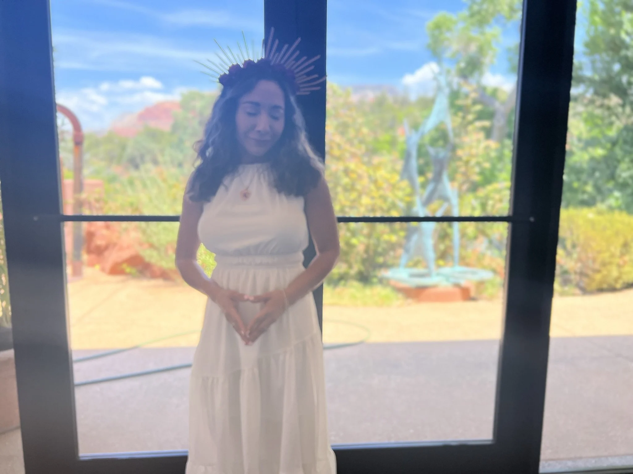 Stephanie in her white dress and ceremonial headpiece beside large windows at her Sister of the Rose initiation