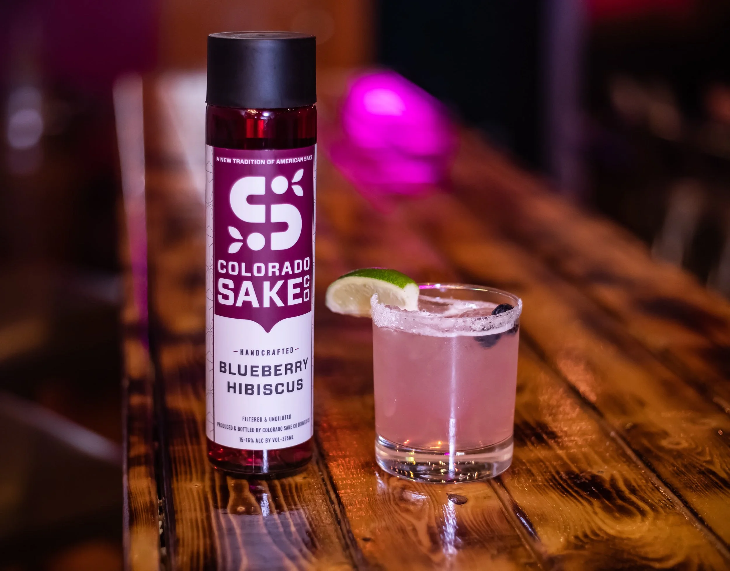 Colorado Sake Co. Blueberry Hibiscus bottle beside a berry cocktail on a taproom table.