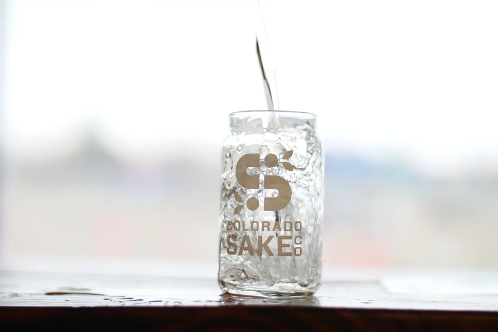 A clear glass with the 'Colorado Sake Co.' logo is being filled with a clear liquid, creating splashes and bubbles, set against a bright, out-of-focus background