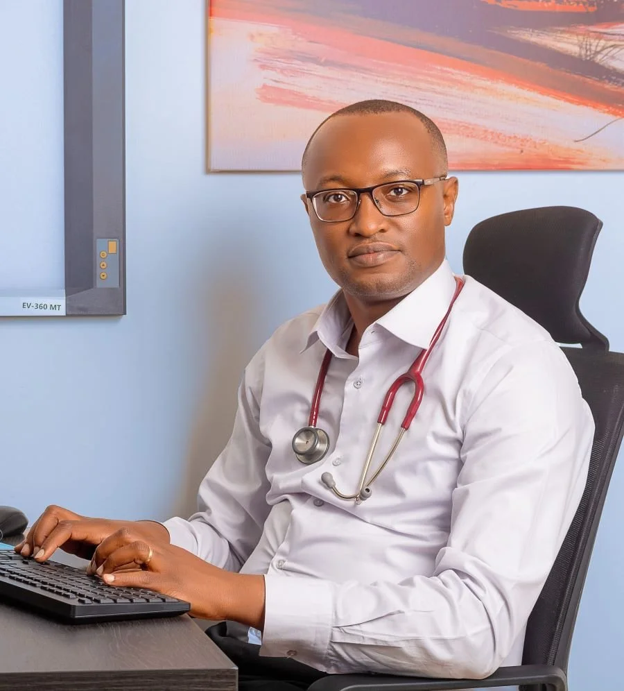 Dr. Timothy Murithi Mwiti seated at a desk with a stethoscope, looking at the camera while working on a computer.
