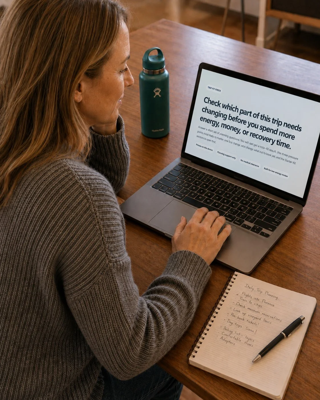 Adult woman in a gray knit sweater sits at a wooden table in a warm home setting, looking at a laptop displaying a Ticked Bucket List Trip Fit Check page. A teal water bottle stands beside the laptop, and an open spiral notebook with handwritten Italy trip-planning notes and a pen rests on the table.