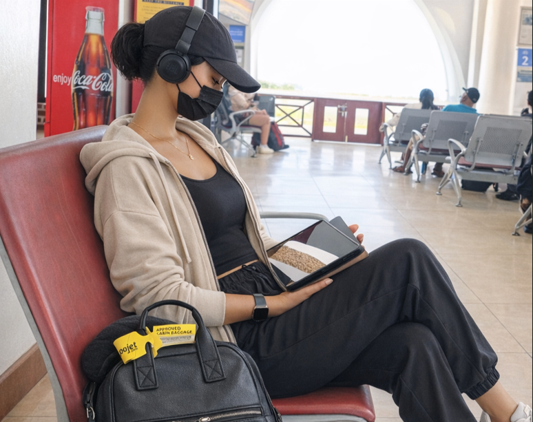 Traveller sitting in an airport lounge with a cleared carry-on luggage waiting to board while reviewing a simple travel plan on a tablet.