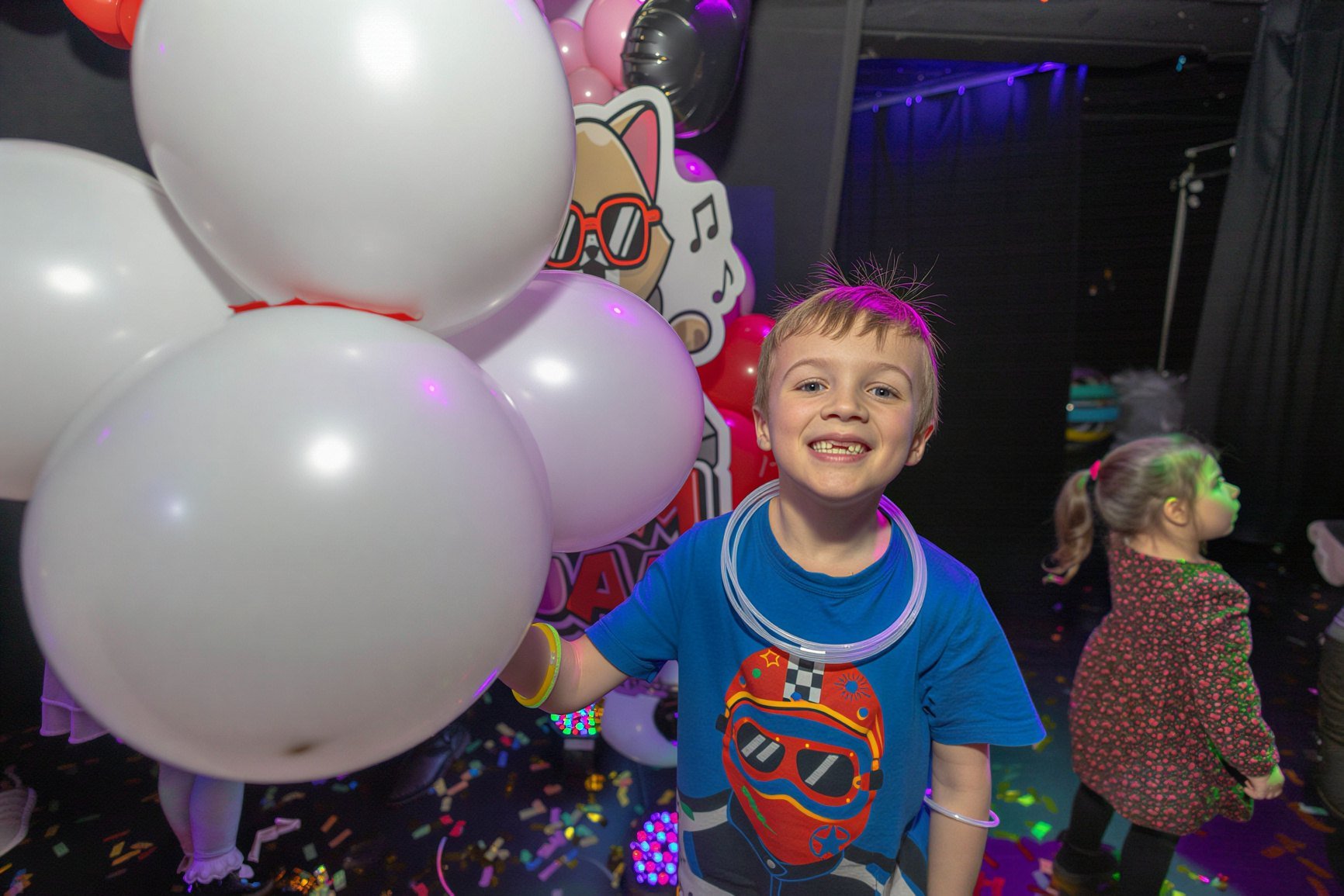 A smiling boy with a glow necklace holding balloons at Fam Jam