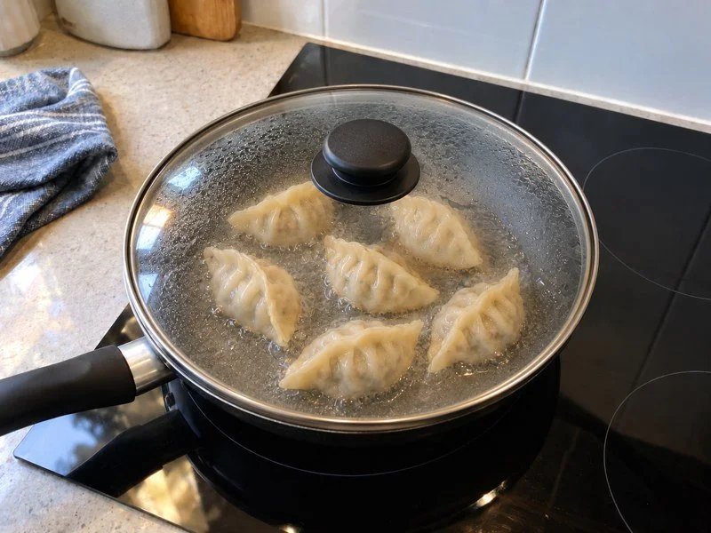 Dumplings cooking in shallow water under the frying pan’s glass lid. / 饺子在平底锅的浅水中煮制，上面盖着玻璃锅盖。