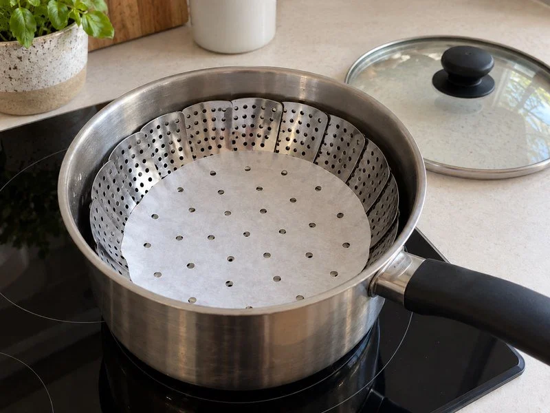 An everyday saucepan with a collapsible steamer basket lined with perforated parchment. / 家用锅内放着折叠蒸篮，铺有带孔蒸垫。