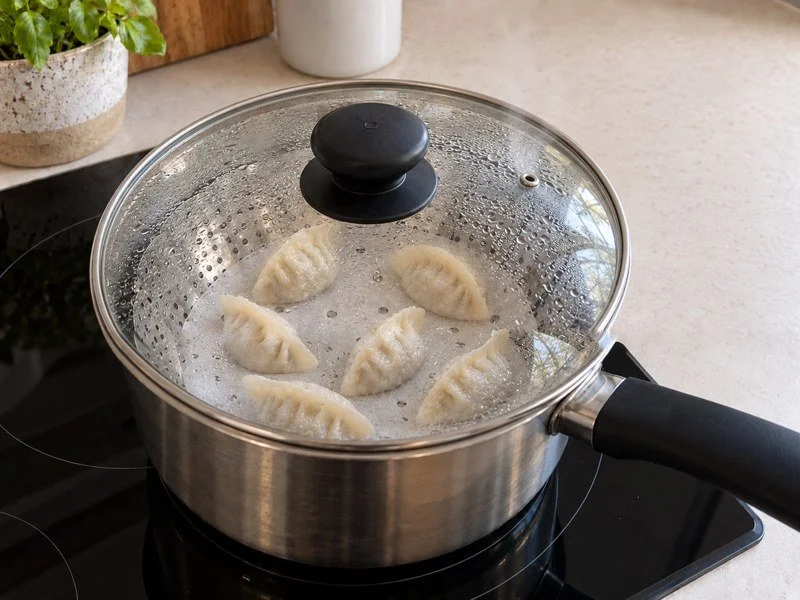 Dumplings steaming on the lined rack beneath a condensation-covered glass lid. / 饺子在铺有蒸垫的蒸屉中蒸煮，玻璃锅盖上凝着水珠。