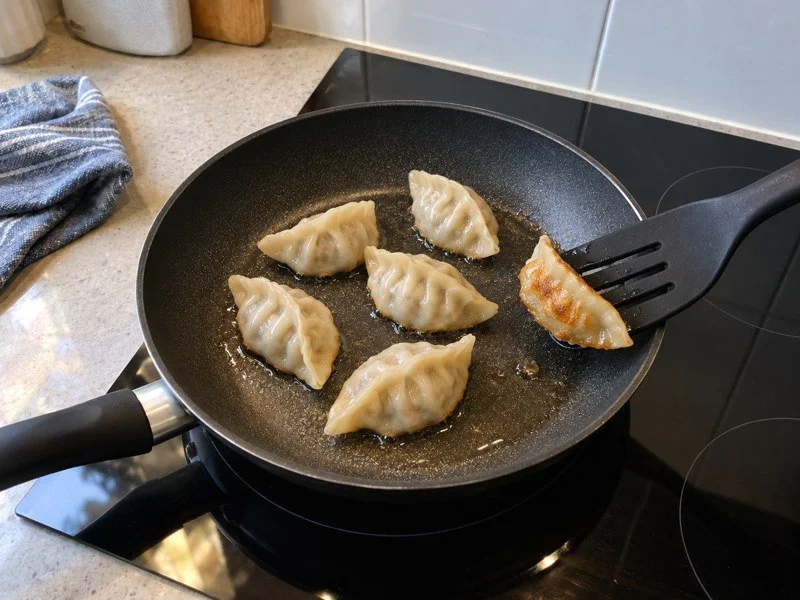 Dumplings frying in a non-stick pan, with one tilted to show its golden underside. / 饺子在不粘锅中煎制，其中一只被翻起，露出金黄底面。