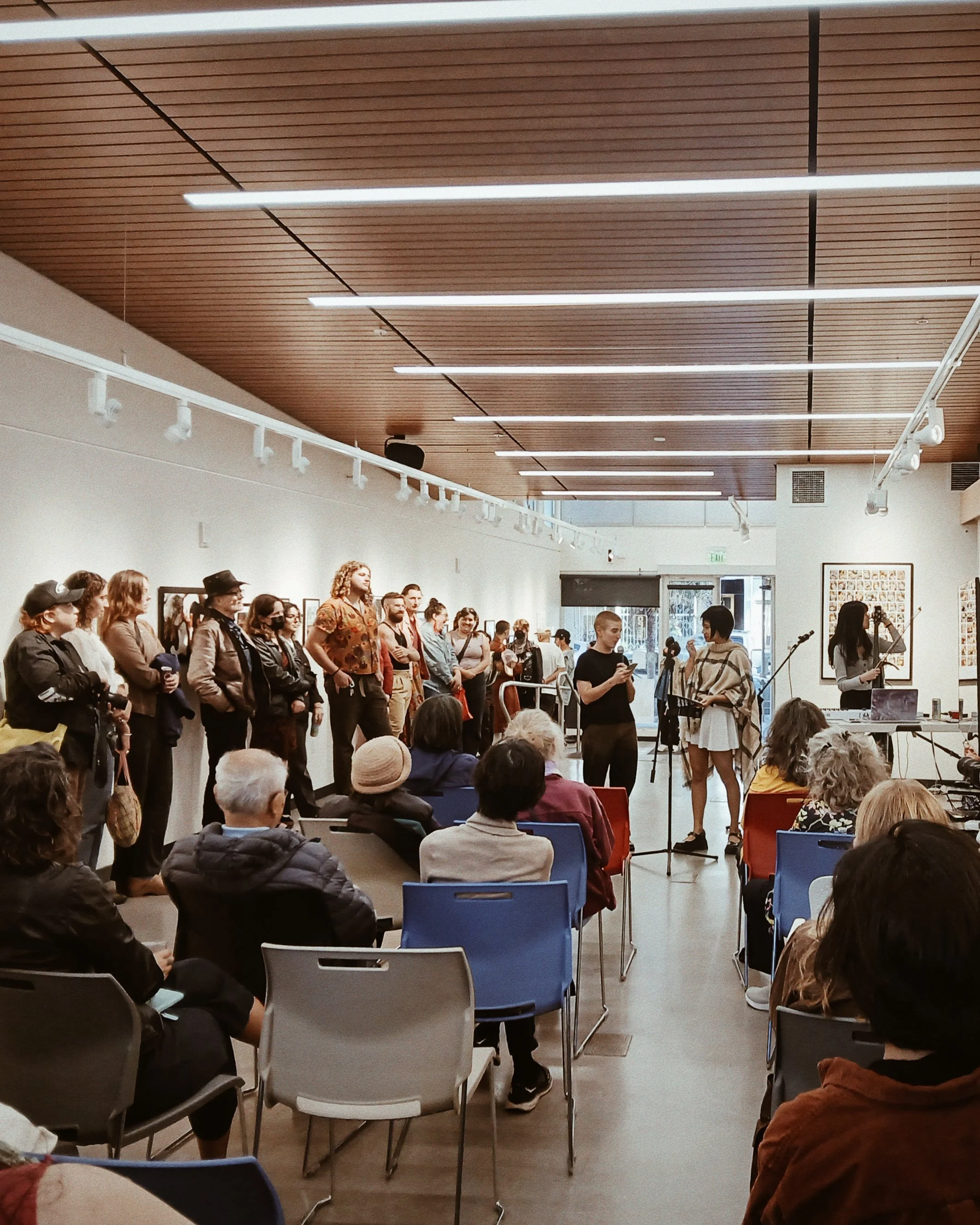 A poet, musician, and exhibition curator stand, speak, and perform in front of a room of sitting and standing people in a small art gallery.