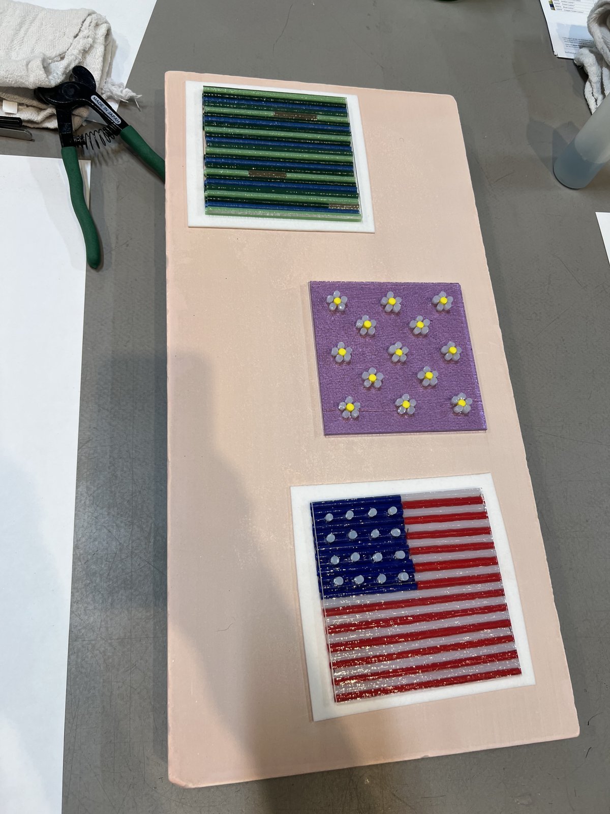 Three fused glass art projects on a work board: green and blue stripes, purple daisies, and an American flag