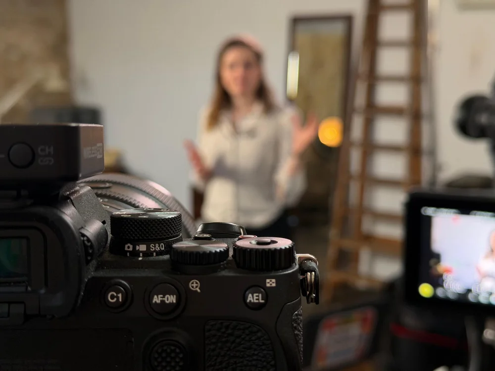 A camera in sharp focus in the foreground while Jocelyn Burnham presents behind it during an Access Learning filming day, 2025