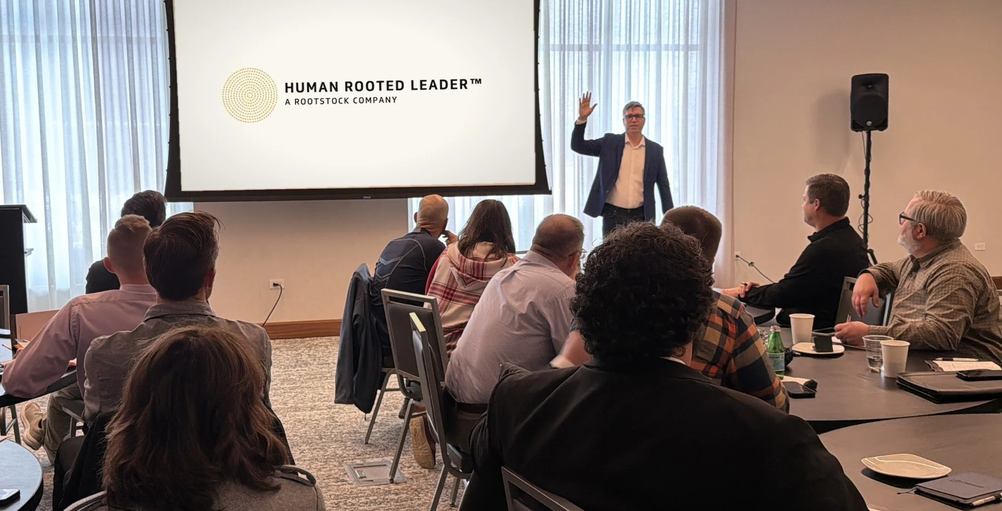 A presenter at the front of a workshop room speaking to seated leaders, with the Human Rooted Leader logo on the screen behind him