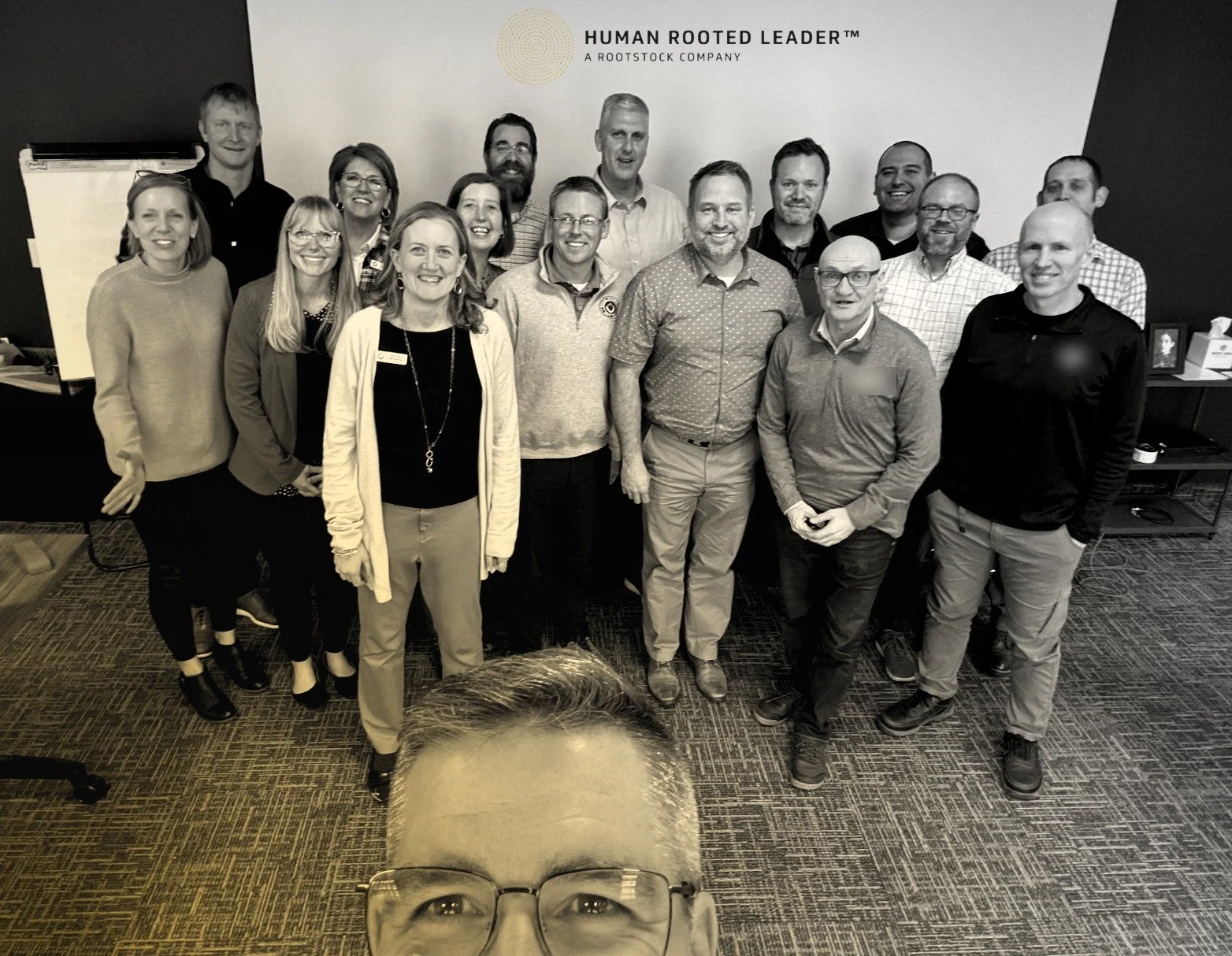 About fifteen leaders smiling together in a meeting room, with the Human Rooted Leader logo on the wall behind them and the photographer’s face in the foreground