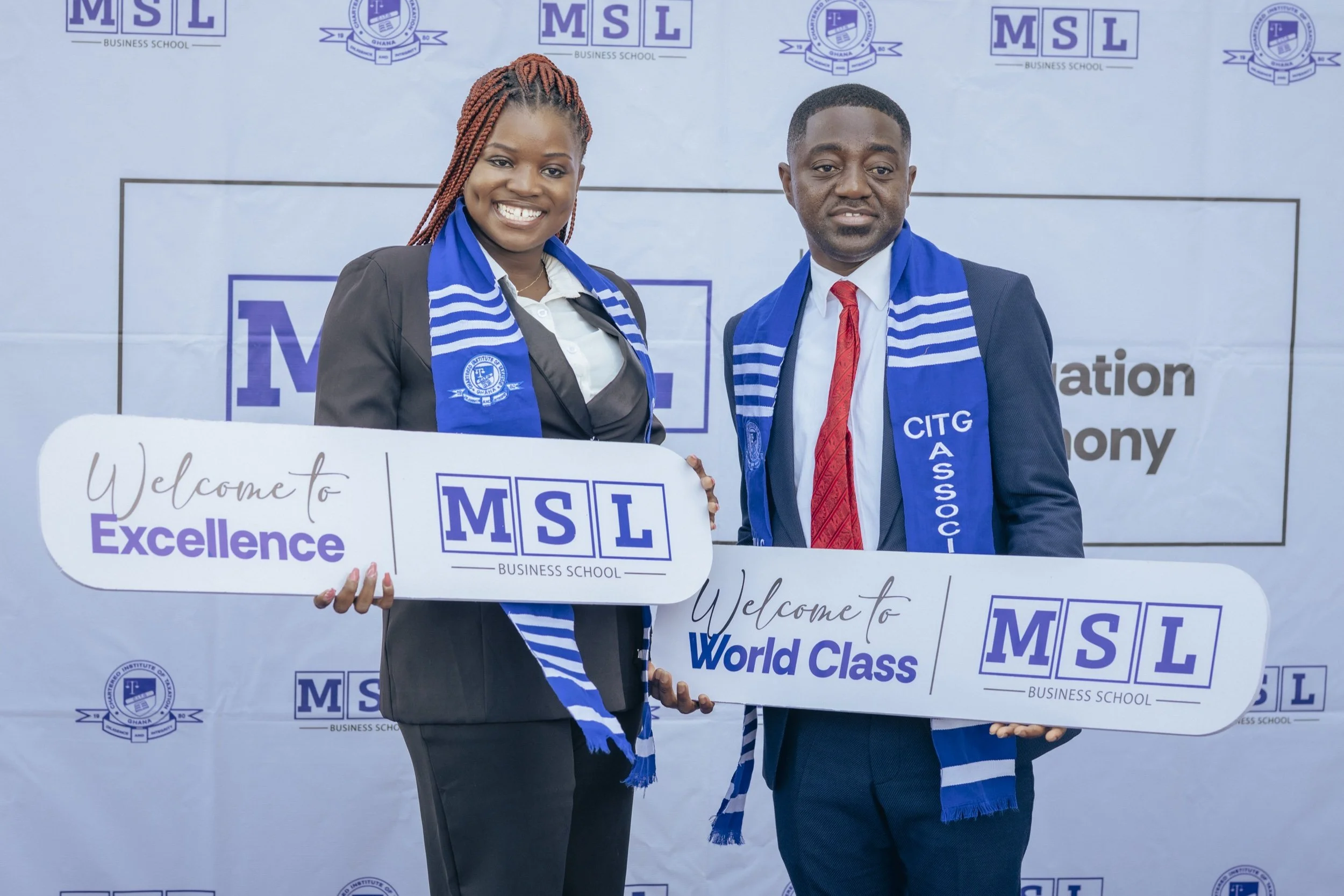 Two MSL students wearing CITG scarves and holding Welcome to Excellence and Welcome to World Class placards in front of an MSL backdrop