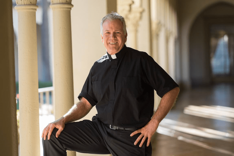 Fr. Jim Clarke, smiling, in a black clerical shirt and collar, in a sunlit arched colonnade.