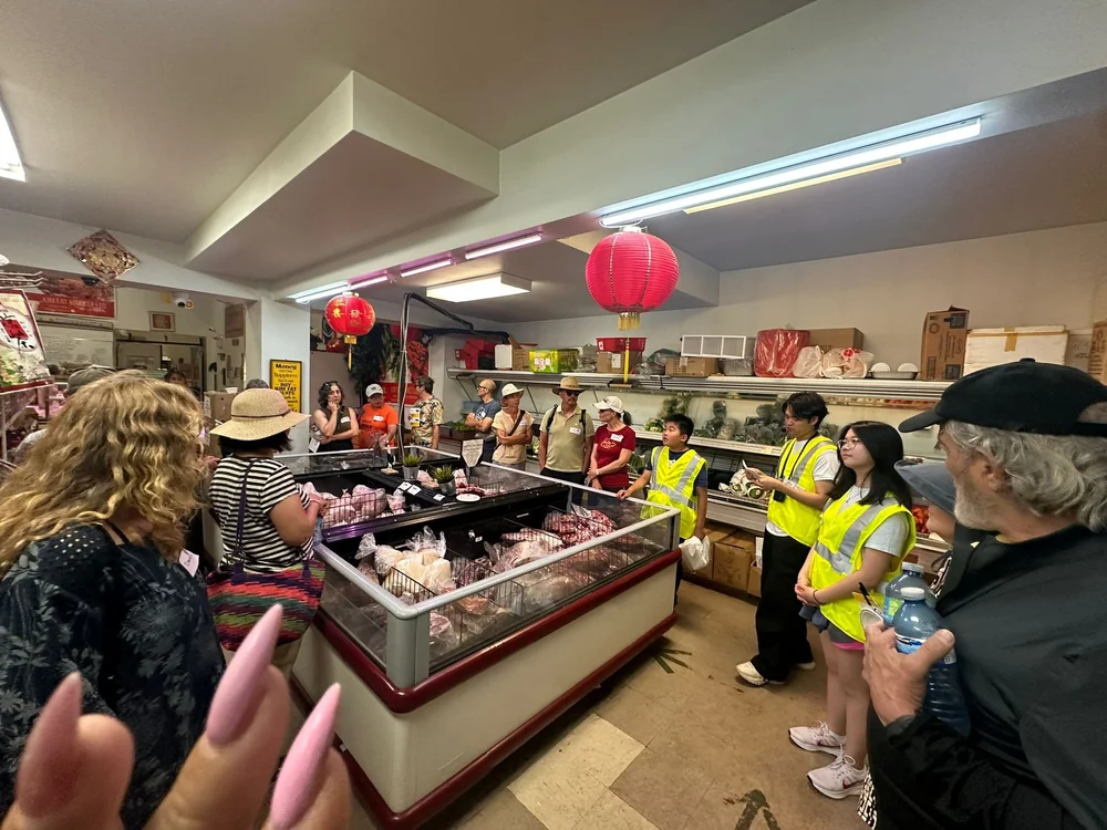 Youth guides in yellow vests lead a Dragon Food Tour group through a Chinatown grocery store.