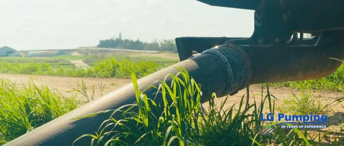 Large dragline hose connected to pumping equipment beside grass, with a farm lagoon in the background.