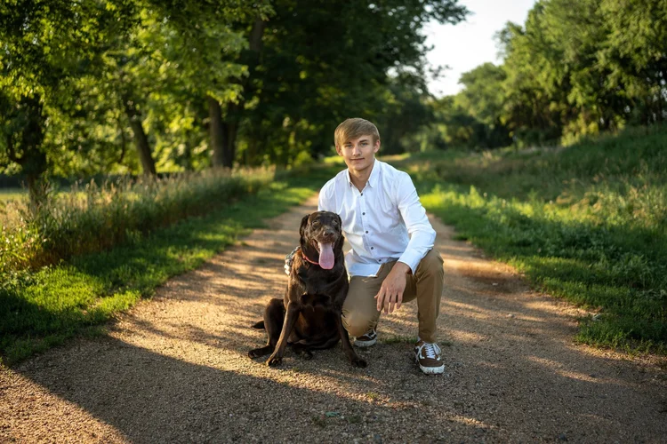 Gavin kneeling beside his chocolate Labrador on a tree-lined gravel road