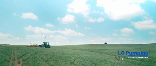 Wide Nebraska crop field with a distant tractor and applicator, wind turbines and a blue sky with clouds.