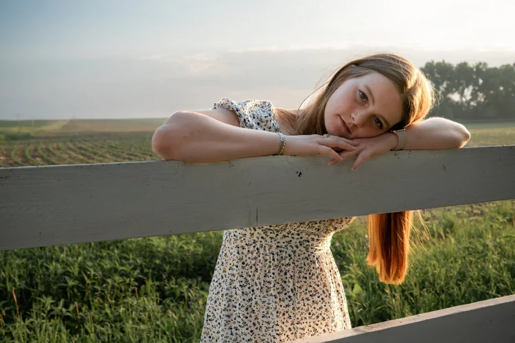 Bella resting her arms on a white wooden fence with open fields behind her