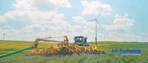 John Deere tractor pulling yellow manure application equipment across a Nebraska crop field, with dragline hoses and wind turbines.
