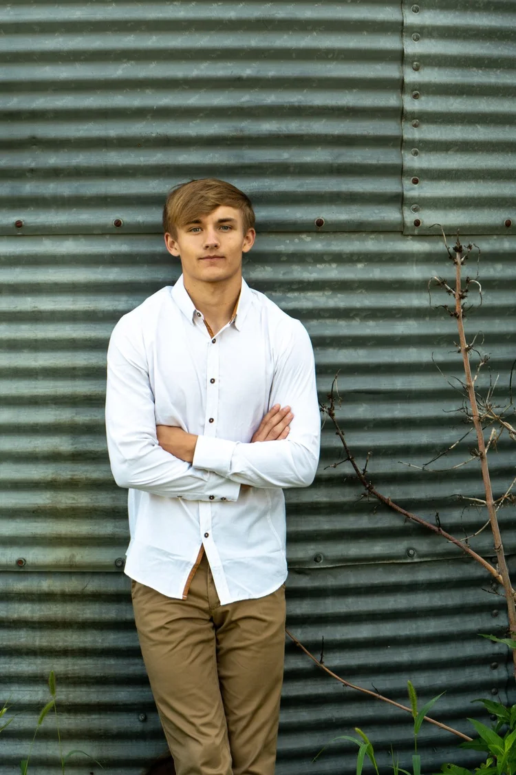 Gavin standing with his arms crossed in a white shirt beside a corrugated metal wall