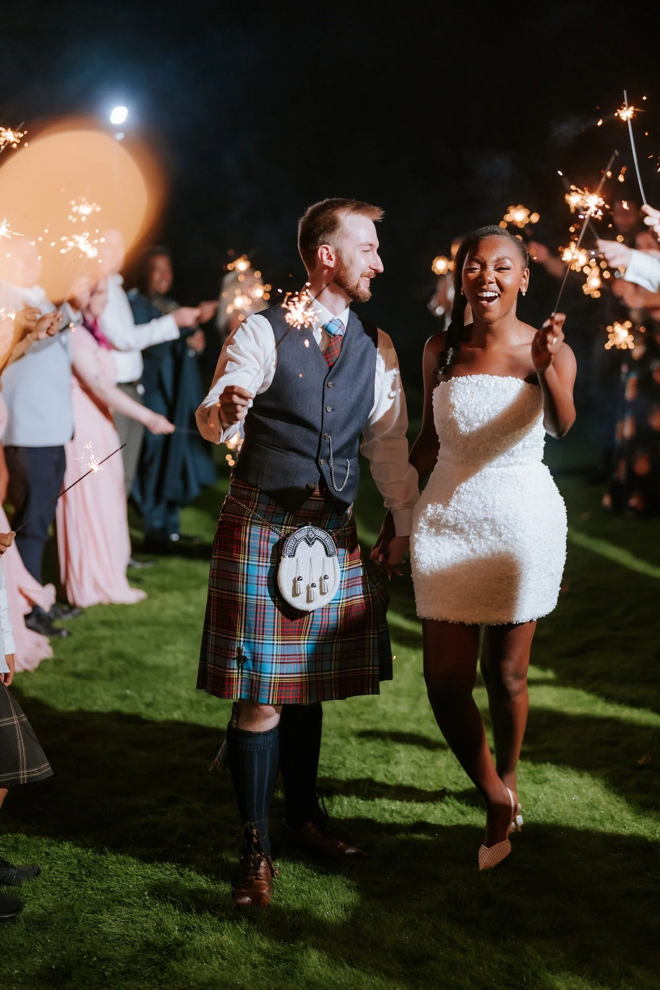 Kuda and Jamie laughing as they walk between guests holding sparklers.