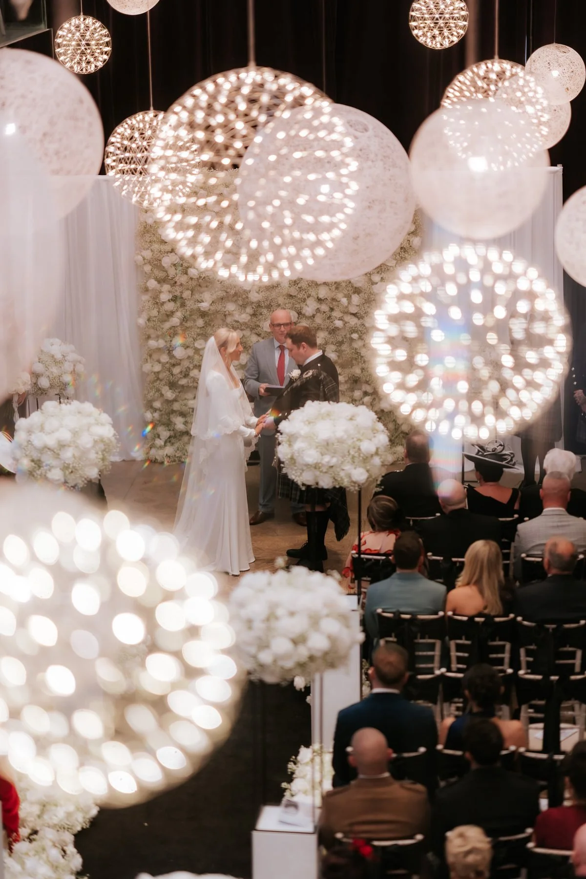 The wedding ceremony framed through the Corinthian’s illuminated hanging globes.