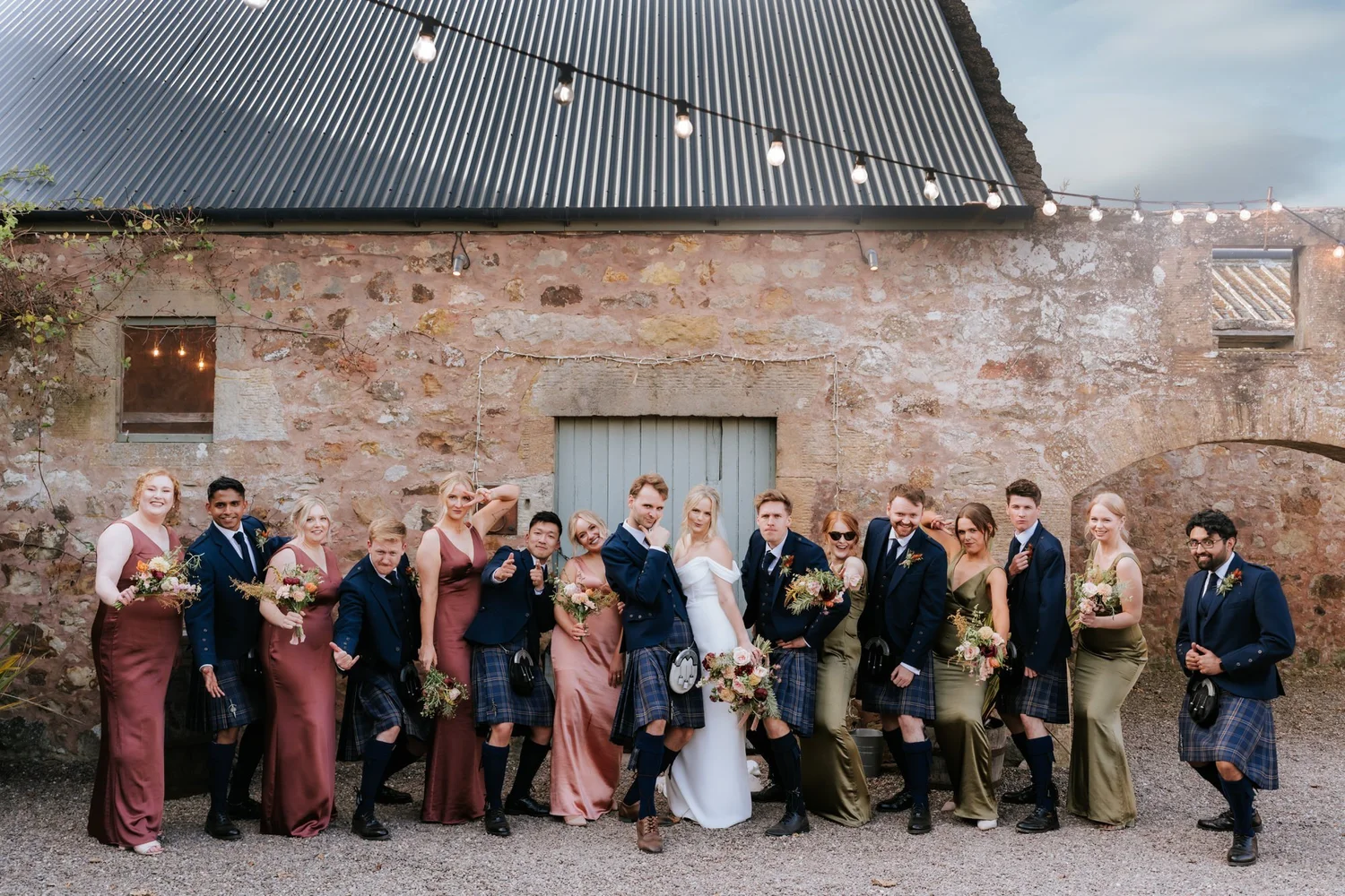 The couple pose with a large group against the venue’s stone wall beneath festoon lights.