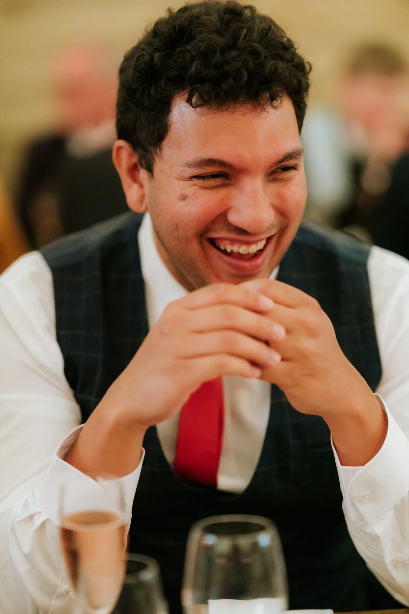 A guest in a checked waistcoat and red tie laughs with his hands clasped beneath his chin.